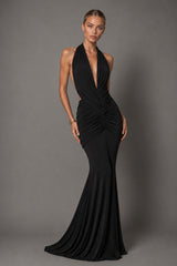 A woman modeling a floor-length black halter gown with a deep plunging neckline, ruched waist, and flared skirt against a plain gray studio background.