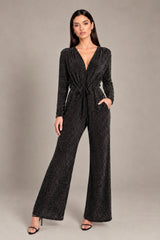 A woman modeling a dark gray sparkly long-sleeve jumpsuit with a deep V-neck, tie waist, and wide legs, standing against a neutral gray studio background.