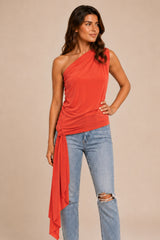 A woman modeling a orange coral red one-shoulder wrap top with distressed light-wash blue jeans, standing against a solid beige studio background.