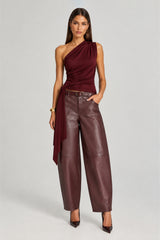 A model stands against a light gray studio background wearing a sleeveless, one-shoulder draped top in deep burgundy and wide-leg faux-leather pants in a matching dark burgundy tone, styled with a thin belt and pointed-toe heels.