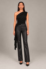 A model wearing a black one-shoulder wrap top and black faux-leather wide-leg pants, standing against a neutral gray studio background.