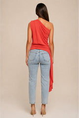 the back of a woman modeling a coral red orange  one-shoulder wrap top with distressed light-wash blue jeans, standing against a solid beige studio background.
