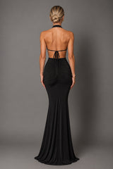 A woman viewed from the back wearing a black halter-neck floor-length gown with a cutout back and gathered ruching, photographed against a plain gray studio background.