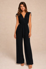A woman modeling a black sleeveless wrap-style jumpsuit with tie details at the shoulders and wide legs, standing against a plain beige studio background.