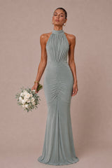 A woman modeling a floor-length sage green halter gown with ruched detailing, holding a white floral bouquet against a solid dusty pink studio background.