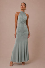 A woman modeling a sleeveless halter-neck maxi dress in a dusty sage green color, standing against a solid warm beige studio backdrop.
