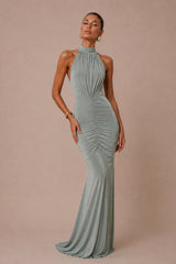 A woman modeling a floor-length sage green halter gown with ruched detailing, standing against a solid beige studio backdrop.