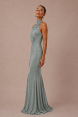 A woman modeling a floor-length, high-neck sage green gown against a solid dusty pink studio background.