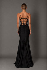 A woman stands with her back to the camera wearing a floor-length black evening gown with thin straps, a fitted silhouette, and an open lace-up back design, photographed against a plain gray studio backdrop.