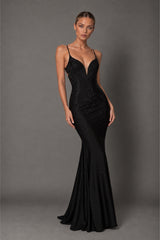 A woman modeling a floor-length black evening gown with thin straps, a deep V-neckline, and subtle beaded embellishments against a solid gray studio background.