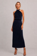 A woman modeling a sleeveless navy blue halter-neck maxi dress with a pleated texture, paired with black strappy high-heeled sandals, against a neutral gray studio background.