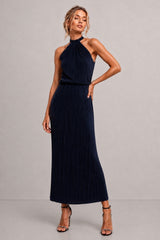 A woman modeling a sleeveless navy blue halter-neck maxi dress with a pleated skirt, standing against a neutral gray studio background.