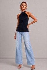 A model wearing a dark navy blue halter top with a textured, pleated surface and light-wash wide-leg denim jeans, standing against a neutral gray studio background.