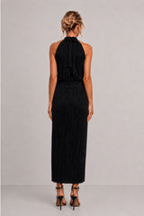 A woman viewed from the back wearing a sleeveless black halter-neck maxi dress with a pleated skirt, paired with black high-heeled sandals, against a neutral light gray studio background.