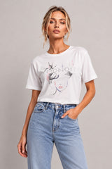 A woman modeling a white graphic t-shirt with a stylized face and the word 'HONOR' printed on the front, paired with light-wash blue jeans, against a solid light gray studio background.