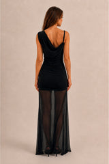 A woman wearing a black sleeveless maxi dress with a draped cowl neckline, ruched bodice, and a sheer floor-length overlay skirt, shown from the back against a plain beige studio background.
