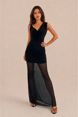 A woman modeling a black sleeveless maxi dress with a draped cowl neckline, ruched bodice, and sheer black tights with a long flowing overlay skirt, posed against a solid beige studio background.