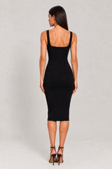 A woman viewed from the back wearing a fitted black midi dress with wide shoulder straps and black strappy high heels, standing against a light gray studio background.