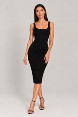 A woman modeling a fitted black sleeveless midi dress with a square neckline and structured seams, paired with black strappy high-heeled sandals, against a light gray studio background.