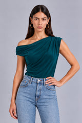 A woman modeling a dark teal green off-the-shoulder draped top paired with light-wash blue jeans against a solid gray studio background.