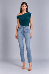 A woman modeling a dark teal green one-shoulder draped top paired with light-wash blue jeans and gold high-heeled sandals, standing against a neutral gray studio background.