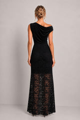 A woman viewed from the back wearing a black off-the-shoulder maxi dress with a velvet bodice and a sheer lace lower skirt, photographed against a plain light gray studio background.
