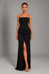 A woman modeling a floor-length black gown with thin straps, a draped ruched bodice, and a high front slit, photographed against a plain gray studio background.