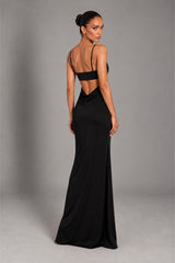A woman modeling a floor-length black gown from the back, featuring thin shoulder straps, a structured back, and a draped open back detail, set against a plain gray studio background.
