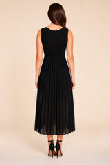 A woman shown from the back wearing a sleeveless black midi dress with a pleated skirt, paired with black high-heeled sandals, against a solid beige studio background.