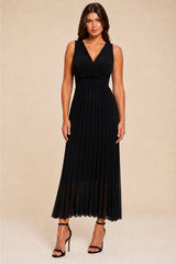 A woman modeling a sleeveless black maxi dress with a deep V-neckline and a pleated skirt, paired with black strappy high-heeled sandals, set against a solid beige studio background.