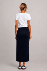 A woman shown from the back wearing a white short-sleeve t-shirt and a dark navy pleated maxi skirt, paired with white sneakers, against a neutral light gray studio background.