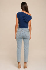 A woman shown from the back wearing a navy blue draped one-shoulder top and light-wash blue jeans, standing against a plain beige studio background.