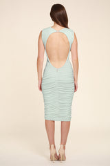 Model wearing a mint green sleeveless midi dress shown from the back. The dress features a dramatic low open-back design with a small button fastening at the neckline, body-contouring ruching through the skirt, and a centre-back seam with gathered detailing. The model wears nude strappy high-heeled sandals and has long, straight brown hair. She is photographed against a soft warm beige studio background.